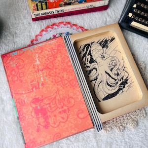 May include: A hollowed-out book with a bright orange cover featuring a decorative pattern. The interior compartment reveals a black and white ink illustration of an octopus. The book spine is accented with black and white vertical stripes.