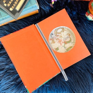 May include: An open book with an orange cover and a circular cutout revealing a colourful illustration of two dragons fighting in the clouds. The book is lying on a blue fluffy surface.