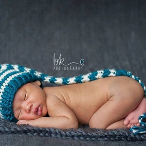 May include: A newborn baby wearing a blue and white striped crocheted hat with a long tail, sleeping on a gray textured blanket.