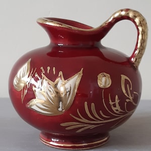 Może przedstawiać: Mały, czerwony dzbanek ceramiczny z pozłacanym uchwytem i kwiatowym wzorem. Dzbanek ma biały i złoty kwiatowy wzór na czerwonym tle.