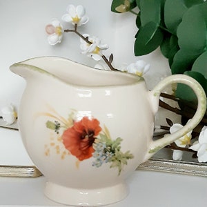 Könnte beinhalten: Ein cremefarbener Keramik-Milchkännchen mit grünem Rand und einem Blumendesign mit einer roten Mohnblume, grünen Blättern und gelben Akzenten. Der Milchkännchen hat einen abgerundeten Körper und einen geschwungenen Griff.