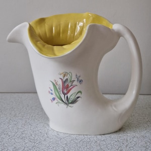 以下が含まれることがあります： 黄色い内側と湾曲したハンドルが付いた白いセラミックピッチャー。ピッチャーには、赤、緑、青のアクセントが付いた花の模様があります。装飾品です。