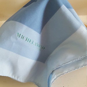 May include: A folded, light blue silk scarf with a geometric pattern of varying shades of blue. The scarf features the name "MICHELANGELO PARIS" in green script. The scarf is made of a soft, flowing material.