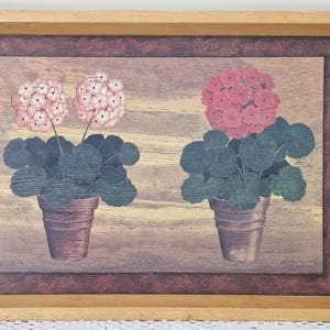 May include: A framed print depicting two potted geraniums with pink and red flowers. The image is set against a wood-effect background, framed by a dark burgundy border and a light wood frame. The artwork has a vintage feel.