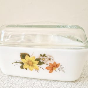 May include: White rectangular casserole dish with a clear glass lid. The dish has a floral design with yellow, orange, and white flowers. A vintage kitchenware item.