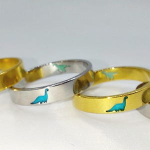 May include: Four rings with dinosaur designs. Two rings are gold with green dinosaurs, and two rings are silver with blue and green dinosaurs.