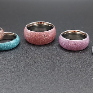 May include: A collection of colorful glitter rings. The rings have a stainless steel interior and come in various colors, including brown, teal, red, purple, silver, and gold. The rings are displayed on a dark surface.