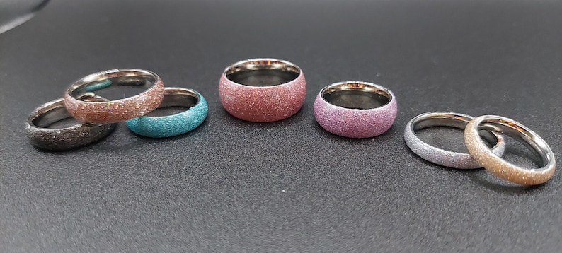 May include: A collection of colorful glitter rings. The rings have a stainless steel interior and come in various colors, including brown, teal, red, purple, silver, and gold. The rings are displayed on a dark surface.