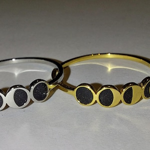 May include: Two rings featuring a moon phase design. One ring is silver-toned with black circular inlays depicting various lunar phases, while the other is gold-toned with matching black circular inlays. Both rings have a thin, polished metal band.