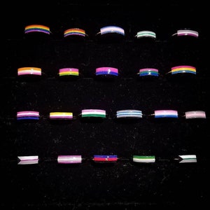 May include: A collection of 18 rings with different colored stripes, representing various pride flags. The rings are arranged in three rows of six, on a black background.
