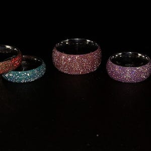 May include: A collection of glittery rings in various colors, including silver, red, teal, pink, and purple. The rings are set against a black background, showcasing their sparkling surfaces. The rings are a wide band style.