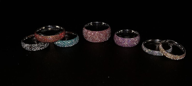 May include: A collection of glittery rings in various colors, including silver, red, teal, pink, and purple. The rings are set against a black background, showcasing their sparkling surfaces. The rings are a wide band style.