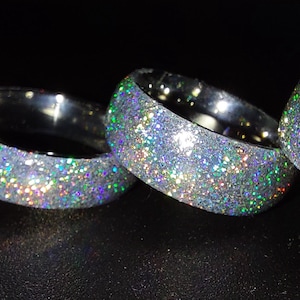 May include: Four silver glitter bangles of varying widths are arranged on a dark surface. The bangles are covered in iridescent glitter, reflecting a rainbow of colors. The bangles are stacked next to each other.