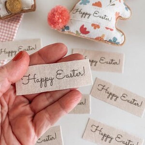 May include: Image of Easter decorations. A hand holds a tag with "Happy Easter!" written on it. Several similar tags are scattered around. Two bunny-shaped ornaments are visible, one with a pom-pom tail. The ornaments are decorated with floral patterns.