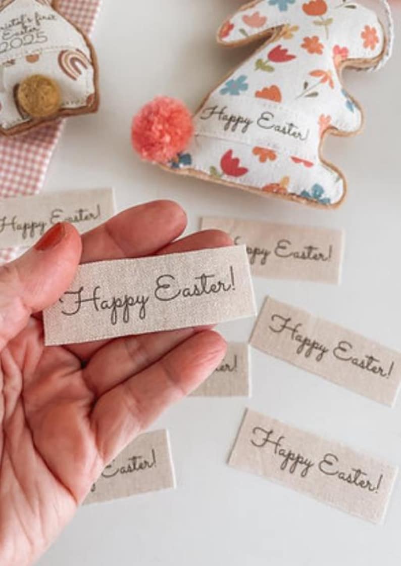 May include: Image of Easter decorations. A hand holds a tag with "Happy Easter!" written on it. Several similar tags are scattered around. Two bunny-shaped ornaments are visible, one with a pom-pom tail. The ornaments are decorated with floral patterns.