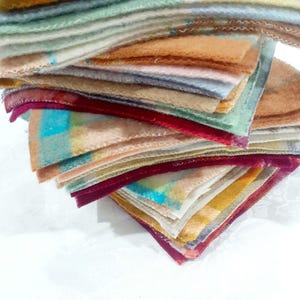 May include: A stack of square fabric swatches featuring a variety of colours including mustard yellow, deep magenta, soft blue, and muted green. Each piece of fabric has a visible zigzag stitched edge, creating a textured and layered appearance.
