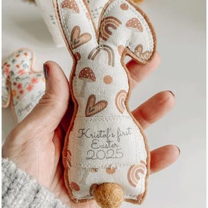 May include: A fabric bunny-shaped Easter decoration with a cream body and brown accents. The bunny has a printed pattern of rainbows, hearts, and other shapes. The text "Kristol's first Easter 2025" is printed on the body. A small, round, brown tail is attached.
