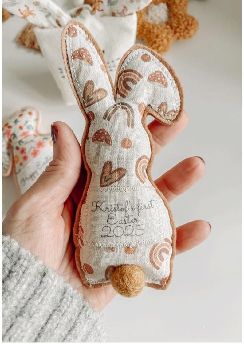 May include: A fabric bunny-shaped Easter decoration with a cream body and brown accents. The bunny has a printed pattern of rainbows, hearts, and other shapes. The text "Kristol's first Easter 2025" is printed on the body. A small, round, brown tail is attached.