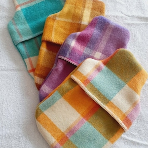 May include: Four hot water bottle covers made of soft, felted fabric, each featuring a distinct plaid pattern in vibrant colours including turquoise, mustard yellow, purple, and cream. Each cover has a visible stitched edge and a small white label.
