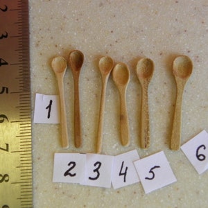 May include: Six small, light-coloured wooden spoons of varying lengths are arranged on a speckled surface. Each spoon has a rounded bowl and a slender handle. Numbers 1 to 6 are written on small white paper tags next to each spoon.