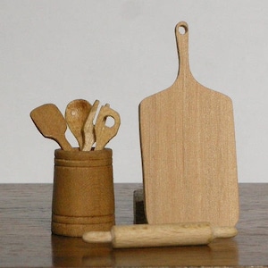 May include: A set of wooden kitchen utensils, including a spoon, spatula, and rolling pin, are displayed in a wooden container. A wooden chopping board with a handle is also included.