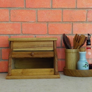 K&ouml;nnte beinhalten: Eine Brotdose aus Holz mit einer Schublade und einem Knopf, neben einem Holzutensilienhalter, einer kleinen blauen Flasche mit floralem Design und einer Glasflasche mit einem Korken. Der Hintergrund ist eine rote Backsteinmauer.