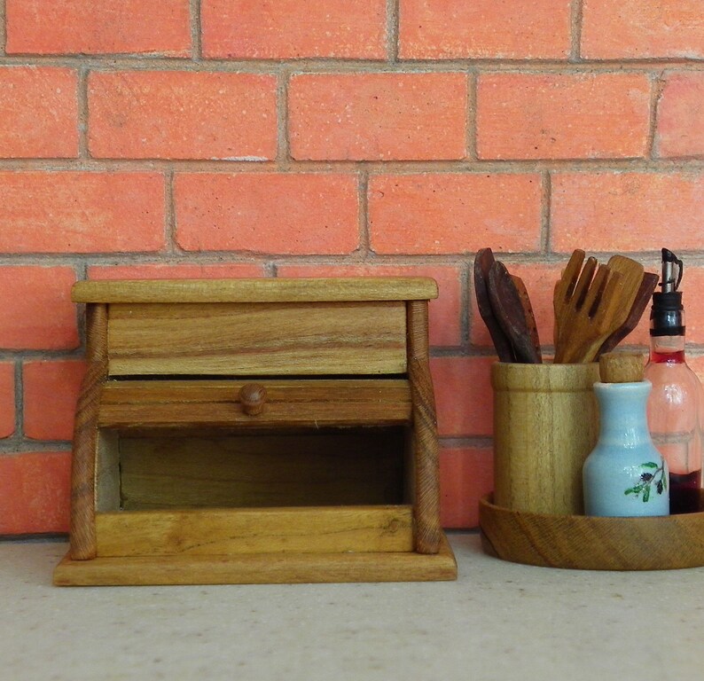 K&ouml;nnte beinhalten: Eine Brotdose aus Holz mit einer Schublade und einem Knopf, neben einem Holzutensilienhalter, einer kleinen blauen Flasche mit floralem Design und einer Glasflasche mit einem Korken. Der Hintergrund ist eine rote Backsteinmauer.
