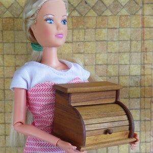 Könnte beinhalten: Eine kleine Brotdose aus Holz mit geschwungener Front und kleinem Knopf. Die Brotdose hat eine warme braune Farbe und scheint handgefertigt zu sein. Die Puppe, die die Brotdose hält, trägt ein weißes Oberteil und ein rosa-weiß gemustertes Kleid.