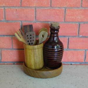 K&ouml;nnte beinhalten: Ein K&uuml;chenutensilien-Set aus Holz mit einer braunen Keramikflasche und einem Korken. Die Utensilien befinden sich in einem hellbraunen Holzhalter, und die Flasche steht auf einem Holztablett. Der Hintergrund ist eine rote Backsteinmauer.