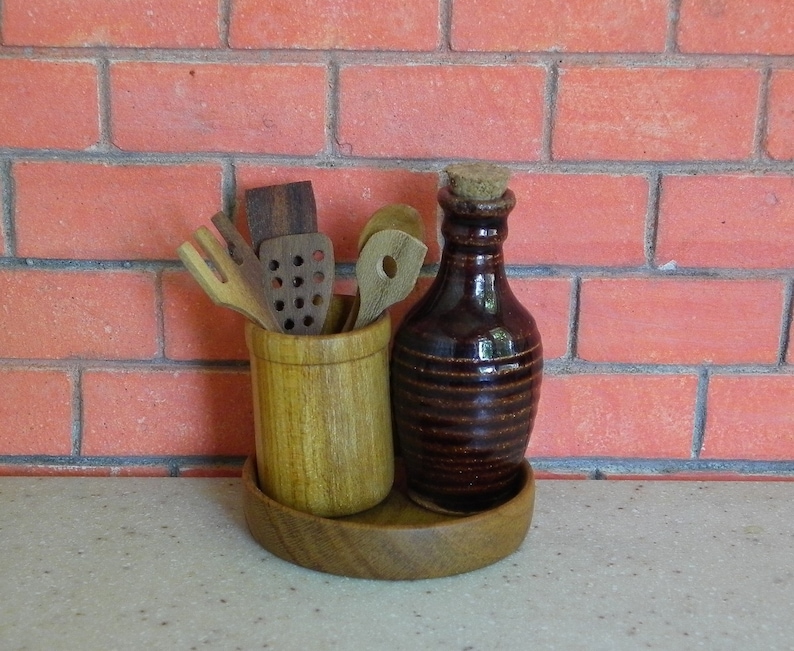 K&ouml;nnte beinhalten: Ein K&uuml;chenutensilien-Set aus Holz mit einer braunen Keramikflasche und einem Korken. Die Utensilien befinden sich in einem hellbraunen Holzhalter, und die Flasche steht auf einem Holztablett. Der Hintergrund ist eine rote Backsteinmauer.