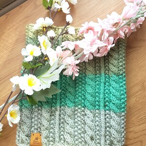 Könnte beinhalten: Ein grüner, grauer und beiger Strickkragen mit einem Zopfmuster. Der Kragen ist mit einem kleinen braunen Lederanhänger verziert, auf dem "Sophie's Knit Stuff" steht.