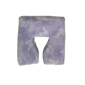 May include: A lavender-colored travel pillow with a white butterfly pattern. The pillow is shaped like a U and has a hole in the middle for the neck.