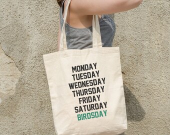 May include: A woman is carrying a beige tote bag with the days of the week printed on it. The last day is misspelled as "Birdsday" in green text.