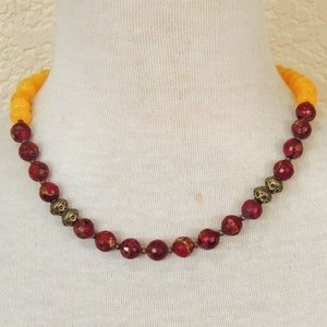 May include: A red and yellow beaded necklace with gold accents. The necklace is made of round beads and is strung on a cord.