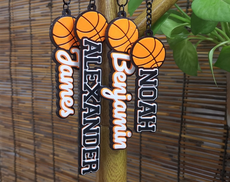 May include: Basketball-themed keychains with personalized names: James, Alexander, Benjamin, and Noah. Each keychain features a basketball design in orange and black, with the name in a contrasting color. The keychains are hanging from a black chain.