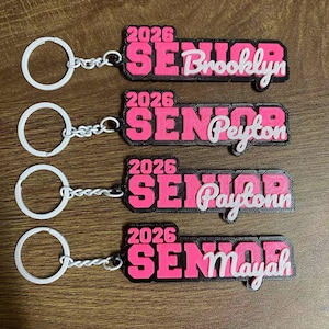 May include: Four keychains with white rings and chains. Each keychain has a black and pink rectangular design with the word "SENIOR" and the year "2026". The names "Brooklyn", "Peyton", "Paytonn", and "Mayah" are written in white.