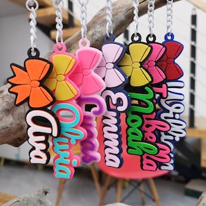 May include: A selection of keyrings with names and bow designs. Each keyring displays a different name in a fun font, accompanied by a bow in various colours, such as orange, pink, yellow, and red. The keyrings are suspended from silver chains.