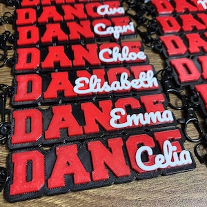 May include: Red and black keychains with the word "DANCE" in bold letters. Each keychain has a different name in white script: Ava, Capri, Chloe, Elisabeth, Emma, and Celia. The keychains have black clasps.