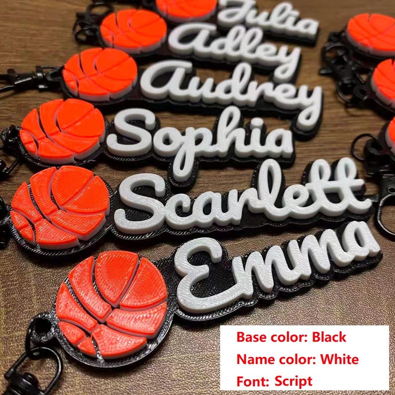 May include: Black keychains with white script names and orange basketball charms. Names include Julia, Adley, Audrey, Sophia, Scarlett, and Emma. The base color is black, the name color is white, and the font is script.