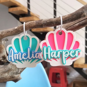 May include: Two white seashell-shaped name tags with teal and pink accents. The name Amelia is on the teal seashell and the name Harper is on the pink seashell.