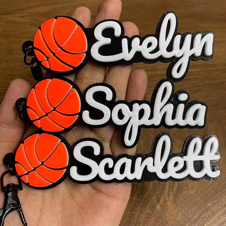 May include: Custom basketball-themed keychains with names Evelyn, Sophia, and Scarlett. Each keychain features a vibrant orange basketball design with black accents and a black clip. The names are in white, with a black outline.