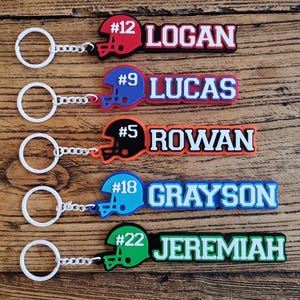 May include: Five personalised football keychain tags with names and numbers. Each tag features a football helmet design in red, blue, black, light blue, and green, with names like Logan, Lucas, Rowan, Grayson, and Jeremiah.