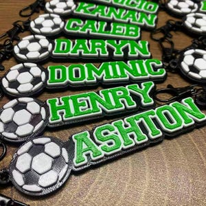 May include: A collection of black and white soccer ball keychains, each featuring a green rectangular name tag with white lettering. The names visible include Kanan, Caleb, Daryn, Dominic, Henry, and Ashton, all attached to black metal clips.