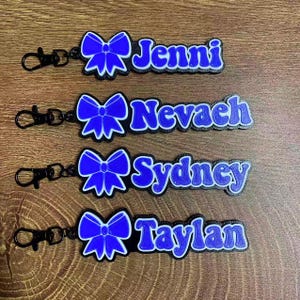 May include: Four personalised keychains with names: Jenni, Nevaeh, Sydney, and Taylan. Each keychain features a blue bow design and a name in blue lettering with a white outline. They have black clasps.
