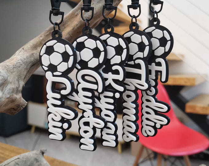 May include: Five black and white silicone keychains featuring a soccer ball at the top and a personalized name in white script lettering. Each keychain includes a black plastic lobster clasp for attachment. The names displayed are Lukin, Ayrton, Kiyaan, Thomas, and Freddie.