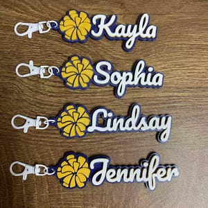 May include: Keychains with names Kayla, Sophia, Lindsay, and Jennifer. Each keychain features a yellow and blue pom-pom design and a white name in a cursive font. The keychains have a silver clasp.