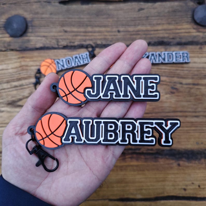 May include: Basketball-themed keychains with personalized names. The keychains feature a basketball design in orange and black, with names like "JANE" and "AUBREY" in bold, white lettering outlined in black. The keychains are attached to black clips.