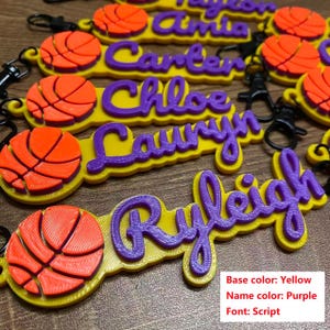 May include: Yellow and purple personalized basketball keychains. Each keychain features a 3D-printed basketball and a name in a script font. The base color is yellow, and the name color is purple.
