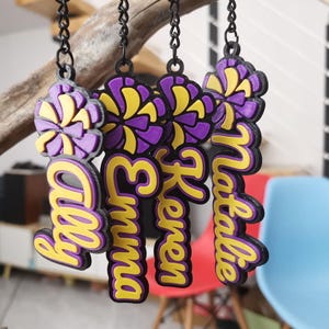 May include: Five purple and yellow 3D printed keyrings with the names Ally, Emma, Keren, Natalie and a purple and yellow pom-pom on top.