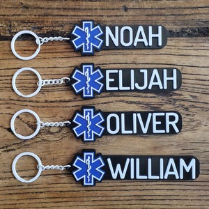 May include: Four black keychains with white text that read "NOAH", "ELIJAH", "OLIVER", and "WILLIAM". Each keychain has a blue and white medical symbol and a silver key ring.